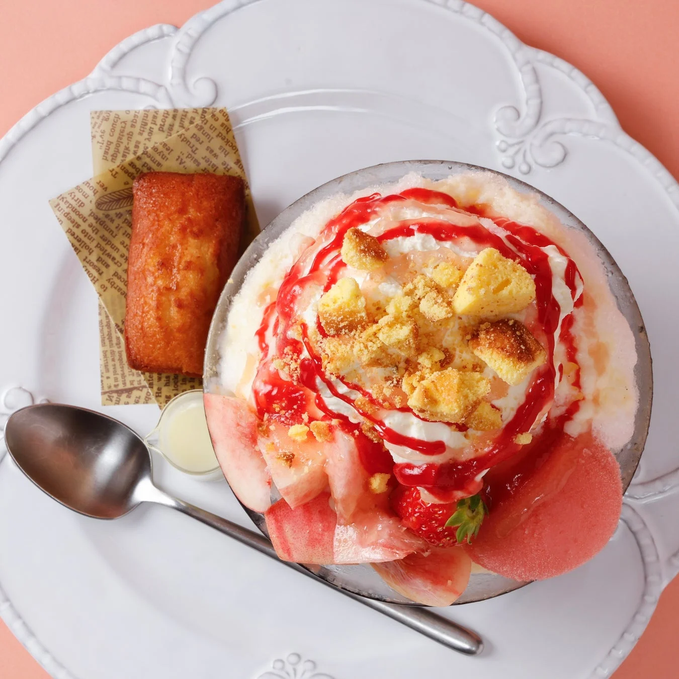 白い皿に盛られた、桃といちご、ホイップクリームがたっぷりのった華やかなかき氷と、フィナンシェが添えられたデザートの俯瞰写真です。夏にぴったりの涼しげで美味しそうな一品です。