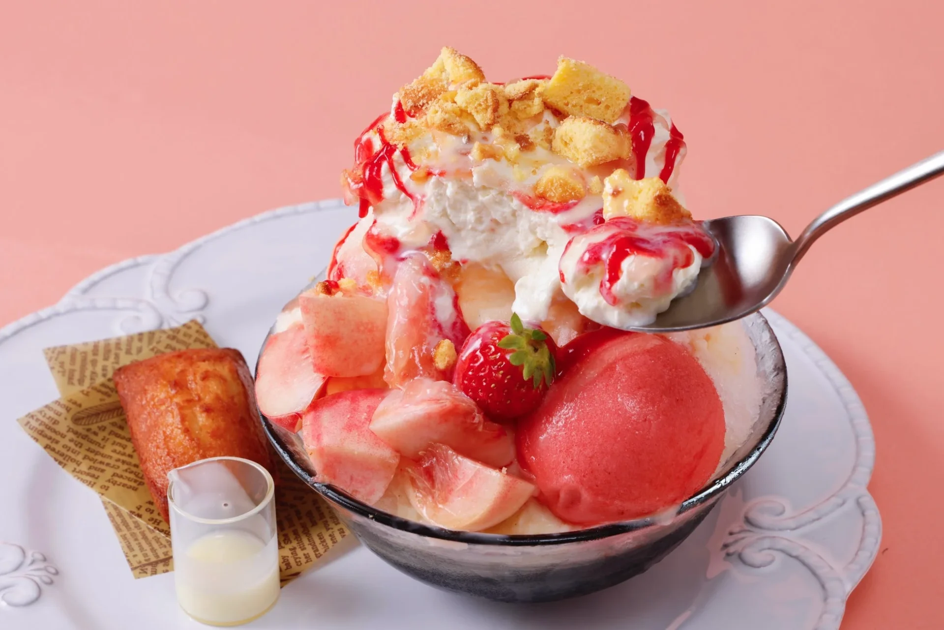 桃、いちご、ホイップクリーム、赤いソース、ケーキクラムがたっぷり乗った豪華なかき氷が、白いお皿にミニパンとミルクピッチャーと共に盛り付けられています。食欲をそそる甘いデザートです。