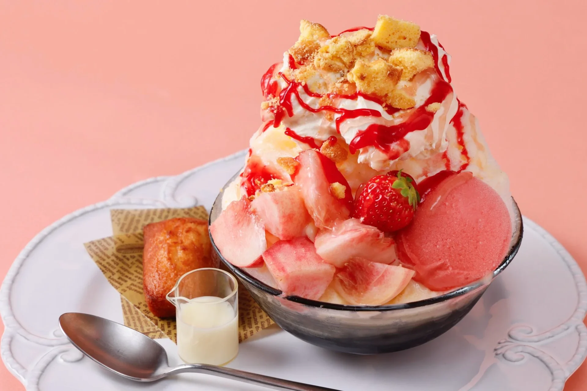 ピンクの背景に、桃やイチゴ、ホイップクリーム、ケーキクラムが飾られた豪華なかき氷が白い皿に盛られています。焼き菓子と練乳が添えられた、見た目も華やかな夏にぴったりの冷たいデザートです。