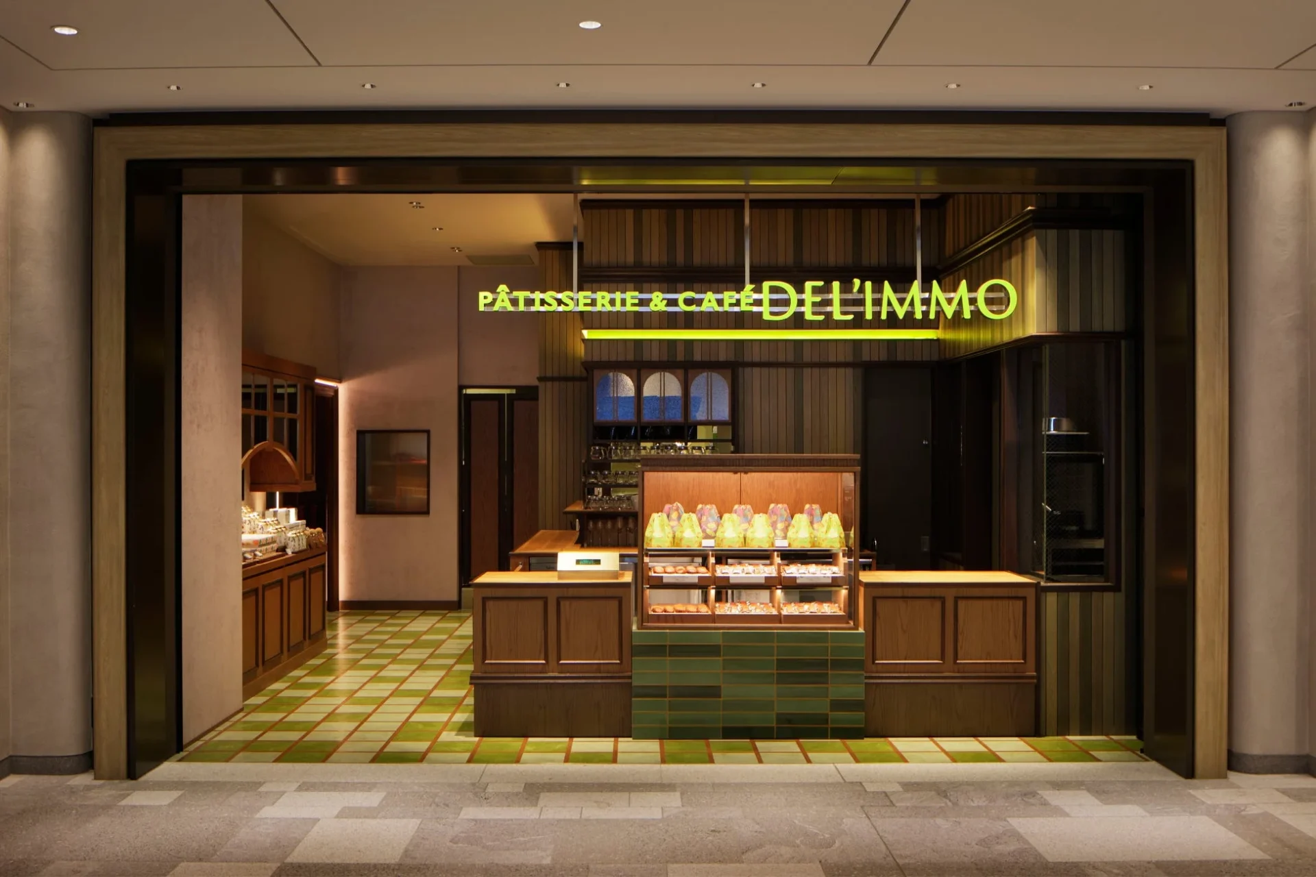 ネオンサインが印象的な「PÂTISSERIE & CAFÉ DEL'IMMO」の店舗エントランスです。木目調の落ち着いた内装と緑の市松模様の床が特徴で、ショーケースには様々な商品が並んでいます。